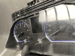 Close-up of a car instrument cluster with tachometer on the left and speedometer on the right; the panel is partially disassembled with two loose probes and a gloved finger in the foreground.