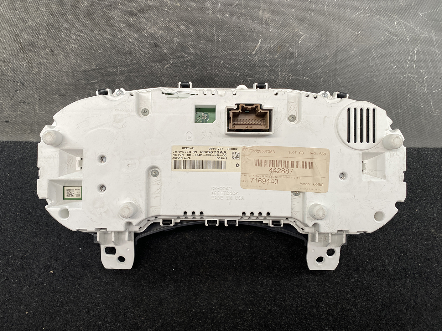 B482_6 Jeep Grand Cherokee WK57A Genuine Instrument Cluster 68235673AA » NZ1.95 » JDM-PARTS NZ » JDM-PARTS NZ Rear view of a car instrument cluster housing in white plastic, with connector ports and a beige label/stickers nearby. Jeep Grand Cherokee WK57A Genuine Instrument Cluster 68235673AA » JDM-PARTS NZ