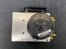 Automotive ABS pump module with a black circular actuator on a metal block and yellow electrical connectors on the side.