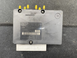 Automotive electronic control unit with yellow LED pins on top, label and barcode on a dark plastic case, mounted on a metal bracket.