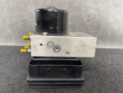 Industrial metal valve block with yellow control knobs on the left, a black cylindrical housing on top, and a black tray underneath on a dark surface.