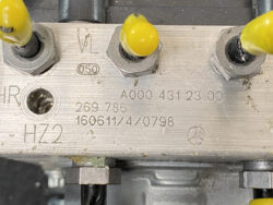 Close-up of a metal valve block with three yellow-tipped terminals and stamped part numbers (A000 431 23 00) and a Mercedes logo, indicating an automotive hydraulic/electrical component.