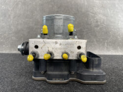 Close-up of an automotive ABS hydraulic valve block with yellow protective caps, mounted on a black base. The metal plate has stamped numbers and fittings are visible on the sides.