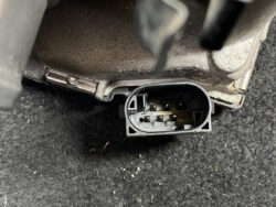 Close-up of a rectangular automotive electrical connector with gold pins, attached to a metal housing.
