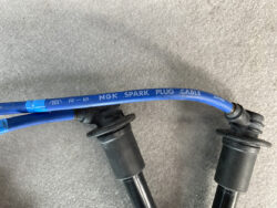 Blue NGK spark plug cable labeled NGK SPARK PLUG CABLE with Japanese text on a gray workshop surface, connecting two ignition components.