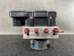 Industrial automotive ABS hydraulic control module with a metal valve block and red test caps, mounted beneath a black plastic electronic unit with labeled ports FL, FR, RR, RL and MC1/MC2.