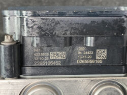 Close-up of a dirty black automotive control module (ECU/ABS unit) with engraved numbers, two QR codes, and a metal mounting bracket below.