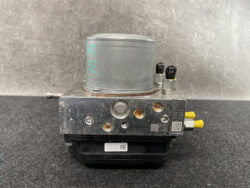 Automobile anti-lock braking system (ABS) pump module with a metal reservoir and electrical connectors, photographed on a dark surface.