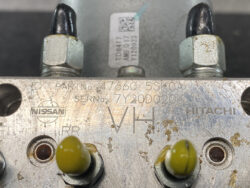 Metal plate with Nissan and Hitachi logos, part numbers, and two yellow-tipped electrical terminals.