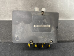 Automotive ABS control module in dusty black plastic with two top connectors, a barcode label, and yellow pins at the bottom on a dark surface.