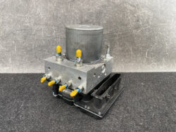 Automotive ABS brake module with yellow-tipped fittings on a metal block and electrical connector on a workshop surface.