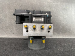 Automotive ABS brake modulator with yellow sensors and a metal valve block, shown on a gray work surface.