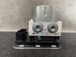 Automotive brake ABS module with two solenoid pistons mounted on a metal block and a black plastic base with a connector on the left side