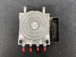 Automotive ABS brake pump module with round motor housing and a multi-pin plug, shown on a dark surface with red-tipped connectors at the bottom.