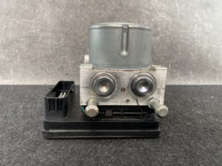 Automotive ABS brake module with a metal valve block and two circular pistons on a black base, left-side electrical connector visible.