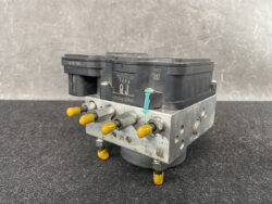 ABS brake module with yellow protective caps on its ports resting on a dark work surface.