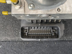 Close-up of a black multi-pin automotive electrical connector plugged into a metal housing, resting on a dark carpeted surface in a vehicle.