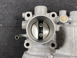 Close-up of a metal throttle body showing a butterfly valve closed inside the circular intake. Two screws secure the valve shaft visible in the center. A small hose barb extends from the side.