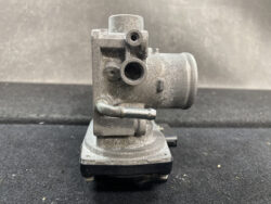 Side view of a dusty, chrome-plated carburetor component with circular intake ports and mounting base, shown on a dark surface.