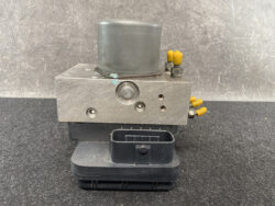 Automotive brake ABS module with metal valve block and yellow electrical connectors on a dark work surface.