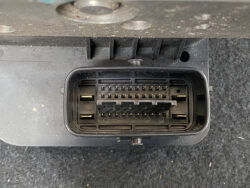 Rectangular automotive electrical connector with two rows of pins and a central locking tab, mounted in a dusty panel.