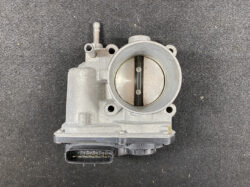Close-up of a car throttle body with two butterfly valves visible inside and an electrical connector at the bottom.