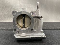 Automotive throttle body with circular intake bore and butterfly valve visible inside, metal housing showing wear and mounting flange.