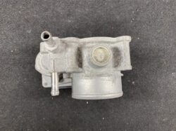Gray metal mechanical component with a protruding tube and a circular plug on dusty surface, viewed from above (likely a hydraulic/valve body).