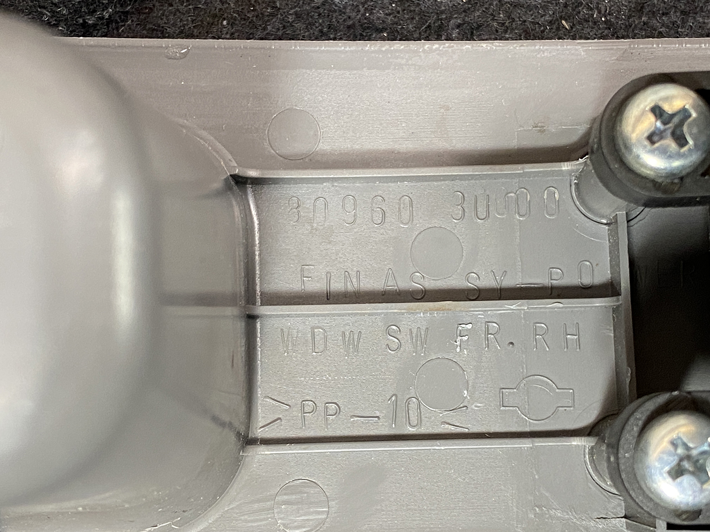 B487_4 NISSAN CUBE Z11 Genuine Master Power Window Switch 80960 3U000 » NZ.95 » JDM-PARTS NZ » JDM-PARTS NZ Close-up of a gray molded plastic panel with embossed alphanumeric codes, circular indentations, and two screws nearby on a dark surface. NISSAN CUBE Z11 Genuine Master Power Window Switch 80960 3U000 » JDM-PARTS NZ