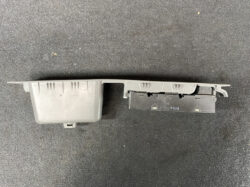 Gray two-piece automotive component with a long plastic housing and a black connector block on a dark carpet surface.