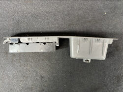 Grey plastic automotive interior trim panel with mounting clips and an attached electrical module, shown on a dark surface.