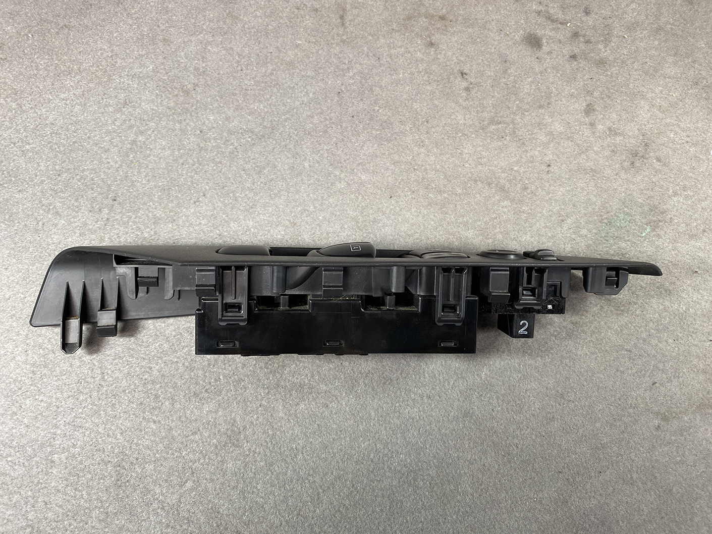 B492_10 NISSAN SERENA SUZUKI LANDY C27 Master Power Window Switch 25401 5TA0A » NZ1.95 » JDM-PARTS NZ » JDM-PARTS NZ Automotive power window switch panel in black plastic, with multiple button housings, shown from the side on a gray surface. NISSAN SERENA SUZUKI LANDY C27 Master Power Window Switch 25401 5TA0A » JDM-PARTS NZ