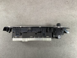 Black automotive switch/console module with multiple mounting tabs and a gray plastic underside, photographed from the side on a gray surface, likely a dashboard control component.