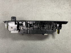 Black automotive control module with a round knob on the top left, showing internal plastic framework and mounting hardware on a gray surface.