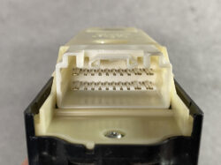 Close-up of a two-row rectangular white electrical connector with many gold pins, mounted on a black housing against a neutral background.