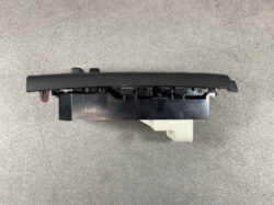 Black textured automotive switch panel with a beige mounting tab, placed on a gray carpet.