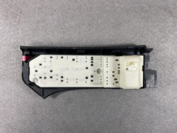 White circuit board inside a black automotive control module, with many small perforations and a right-side connector.