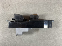 Black automotive electronic module with an orange-ring connector on top, mounted on a gray bracket, viewed from above.
