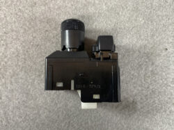 Black automotive switch module with a textured round knob on top and a white mounting tab at the bottom also showing side clips on a gray surface of the background