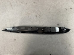 Long black automotive interior trim piece with an integrated switch housing and electrical connector on the right, laying on a gray surface.
