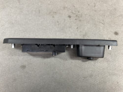 Automotive dashboard control panel mounting unit with side tabs and a right-side knob, placed on a concrete floor.
