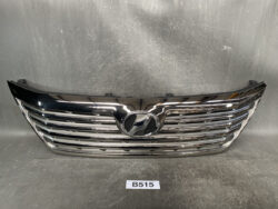 Chrome front grille of a car with a central oval badge, photographed indoors against a scratched gray backdrop