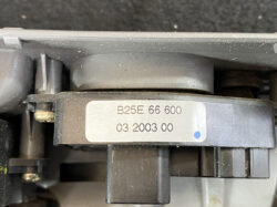 Close-up of a mechanical part with a silver label reading 'B25E 66 600' and '03 2003 00' on a dark plastic bracket, with a small blue dot.