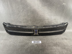 Front grille assembly from a vehicle with a black diamond-mesh pattern and chrome trim, placed on a gray surface with a central emblem visible