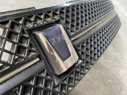 Close-up of a black plastic car grille with a rectangular badge reading VOXAI in the center, surrounded by dusty, textured mesh patterns.