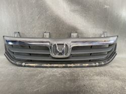 Front grille of a Honda vehicle with a prominent chrome Honda emblem in the center and horizontal black slats on a dusty bumper area.