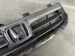 Close-up of a Honda front grille with a chrome emblem on a dark bumper material edge, slight dust present.