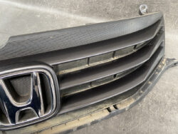 Close-up of a car front grille with a chrome Honda emblem on the left and black horizontal slats behind a dusty bumper
