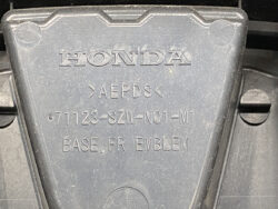 Close-up of a gray plastic Honda emblem base labeled 'BASE, FR EMBLEM' with part number 711123-SZW-N01-M1 on the cover, suggesting it's the front emblem base component.
