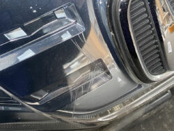 Close-up of a dark blue car bumper with multiple long surface scratches near the front light/grille area in the lower front corner.